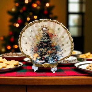 May include: A decorative seashell-shaped plate with a Christmas tree design. The plate has a gold-colored rim and is supported by a clear stand. The Christmas tree is decorated with red ornaments and a pearl-like star.