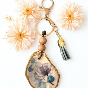 May include: A decorative keychain with an oyster shell pendant, painted with a floral design in blue and purple hues. The shell has a gold trim, wooden beads, and a tassel. The keychain is attached to a gold-coloured clasp.