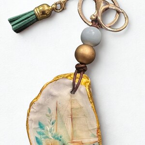 May include: A decorative keychain with a painted seashell design. The shell has a gold-painted edge and features a sailboat image. It includes a green tassel, gold-coloured hardware, and decorative beads. The sailboat image has blue and white accents.