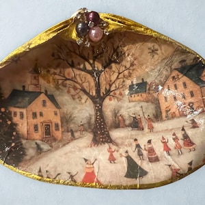 Op de afbeelding: Decoratieve zeeschelp met een winterdorpsscène. De schelp heeft een goudkleurige rand en is versierd met parels en kristallen. De scène toont een besneeuwd landschap met huizen, een kerk en figuren.