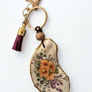 May include: A gold-coloured keyring with a burgundy tassel and a floral pendant. The pendant is an irregular shape with a gold-coloured edge, featuring a bouquet of orange and purple flowers.