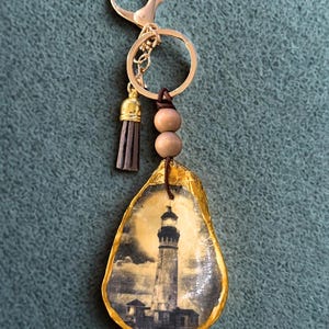 May include: A gold-toned keychain featuring a lighthouse image on an oyster shell. The shell is edged in gold and attached to a gold ring, clasp, and a small tassel. Two wooden beads and a leather cord are also attached.