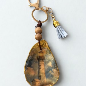 May include: A gold-coloured keychain with an oyster shell pendant. The pendant depicts a lighthouse scene with a gold border, wooden beads, and a grey tassel. The keychain has a lobster clasp and a split ring.