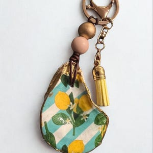 May include: A decorative keychain with a painted oyster shell. The shell features a lemon and leaf design on a blue and white striped background, with a gold edge. It has a gold clasp, yellow tassel, and wooden beads. The keychain is a unique accessory.