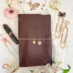 May include: A brown leather journal with a pink elastic band, gold heart charm, and pink heart charm. The image includes a pen, paperclips, binder clips, and a card with a deer illustration and the text "All about that doe."