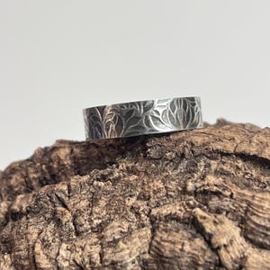 May include: A silver ring with a detailed floral pattern. The ring is set against a textured, brown surface. The band features an engraved design of leaves and vines, creating a nature-inspired aesthetic. The ring's metallic sheen contrasts with the organic background.