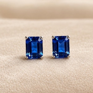 May include: A pair of stud earrings featuring rectangular emerald-cut blue gemstones set in four-prong silver-toned metal settings. The vibrant blue stones have a faceted surface that reflects light, creating a clear and polished appearance.