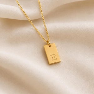 May include: A gold-toned necklace featuring a delicate chain and a rectangular pendant. The pendant has a smooth, polished surface with the capital letter E engraved in the centre. The metal has a warm, reflective finish throughout the entire piece.