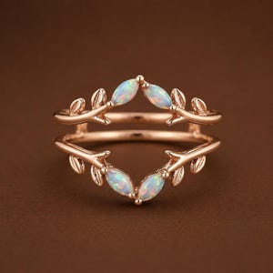 Può includere: Un anello in oro rosa con un design a doppia fascia a forma di rami d'albero con foglie. L'anello è impreziosito da quattro opali taglio marquise che mostrano lampi di colore iridescente, posizionati simmetricamente lungo le fasce metalliche.
