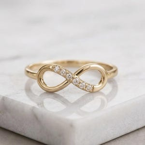 May include: A gold-coloured ring shaped as an infinity symbol. The ring features small, clear gemstones set along the centre of the infinity loop. The ring is displayed on a white marble surface.