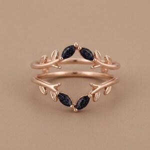 May include: A rose gold ring featuring a double band design shaped like branches with leaves. The ring is accented with four marquise-cut dark blue sandstone gems that have a sparkling, glittery appearance, positioned at the centre of the branches.