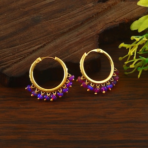 Può includere: Un paio di orecchini a cerchio dorati con una serie di piccole perline rotonde color viola e blu iridescente che pendono dalla metà inferiore di ogni cerchio. Gli orecchini presentano un design circolare con una chiusura a cerniera in alto.