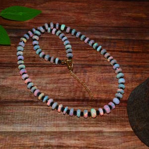 May include: A beaded necklace featuring iridescent opal beads interspersed with black beads. The necklace has a gold-colored clasp and is displayed on a wooden surface. The opal beads display a range of colors.