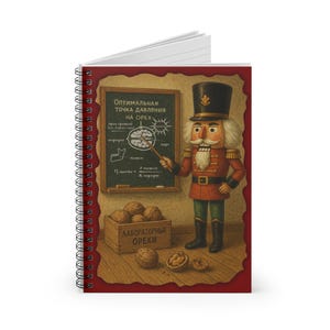 Puede incluir: Cuaderno de espiral con una ilustración caprichosa de un cascanueces con uniforme rojo y dorado. El cascanueces está frente a una pizarra con texto y diagramas en ruso sobre nueces. También se muestra una caja de madera etiquetada como "Laboratory Nuts".