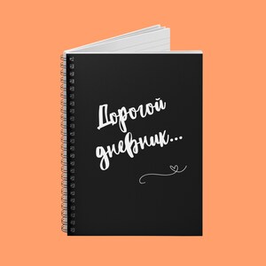 May include: A black spiral-bound notebook with the text "Дорогой дневник..." in white script. The notebook is open, revealing white lined pages. A small heart-shaped design is below the text. The background is a solid orange color.