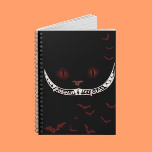 May include: Black spiral-bound notebook featuring a Cheshire Cat design with red eyes and a wide white grin. The notebook cover has the Russian text "рабочая тетрадь" and includes illustrations of bats.