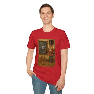 Peut inclure: T-shirt rouge avec une illustration vintage d'un casse-noisette en uniforme rouge et vert, devant un tableau noir avec du texte. Le casse-noisette tient un pointeur. Une caisse en bois avec des oranges est visible.