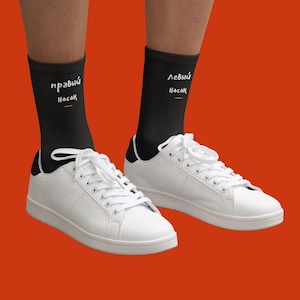 Puede incluir: Zapatillas blancas con detalles negros y cordones blancos, combinadas con calcetines negros. Los calcetines tienen texto cirílico blanco que dice "calcetín derecho" y "calcetín izquierdo". El fondo es naranja.