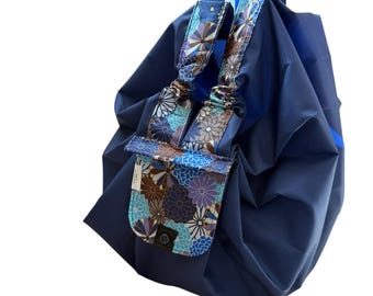 May include: A navy blue tote bag featuring a gathered, rounded silhouette and a front flap pocket. The flap and handle accents are decorated with a multi-colored floral pattern in shades of purple, blue, brown, and white, secured by a black snap.