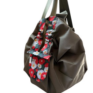 May include: Olive green tote bag with floral accents. The bag features a colorful floral pattern in red, white, and green on the straps and a small pocket. The bag has a simple design with long straps.