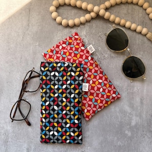 May include: Two rectangular fabric pouches featuring a repeating geometric petal pattern. One pouch has a black background, the other a red background. Both feature a small white tag labeled GEN. Includes brown-rimmed glasses and black sunglasses.