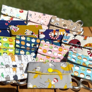 May include: A selection of handmade fabric wallets with diverse patterns, including cats, bananas, and abstract designs. The wallets are in various colours, such as blue, yellow, and pink, and are arranged on a wooden surface.
