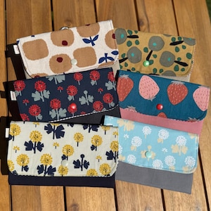 Pode incluir: Uma coleção de carteiras de tecido com vários padrões, incluindo florais, de frutas e abstratos. As carteiras apresentam fechos de pressão e estão em tons de azul, castanho, amarelo e rosa. As carteiras são exibidas numa superfície de madeira.