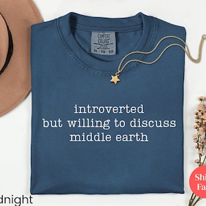 May include: Midnight blue t-shirt with the text "introverted but willing to discuss middle earth." A gold star pendant necklace is on the shirt. A brown hat and flowers are in the background.