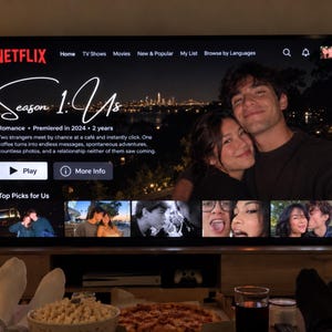 May include: A television screen displaying the Netflix interface for the show Season 1: Us. The screen features a title, a synopsis, a Play button, a More Info button, and a background image of a couple against a city skyline at night.