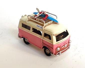 May include: A miniature pink and cream-colored van with a surfboard on top. The van has a luggage rack, black wheels, and the text "2404D-5581" on the roof. The surfboard is blue and white with red accents.