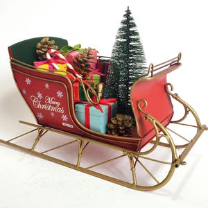 Könnte beinhalten: Ein roter Weihnachts-Metallschlitten mit goldfarbenen Kufen und Akzenten. Der Schlitten ist mit verpackten Geschenken, Tannenzapfen, einem kleinen Weihnachtsbaum und Zuckerstangen gefüllt. Auf der Seite steht "Merry Christmas".
