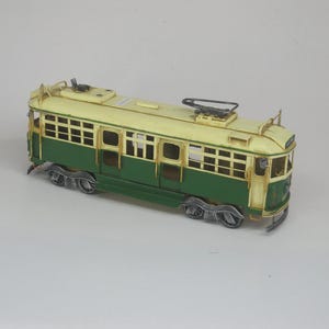May include: A vintage model trolley car in cream and green. The trolley has detailed windows, wheels, and overhead electrical equipment. The model is a decorative item, likely a collectible or a nostalgic piece.