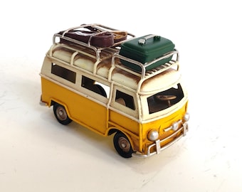 May include: A miniature yellow and white vintage van with a luggage rack on top. The rack holds a green suitcase and two brown containers. The van has black wheels and a detailed front grill.