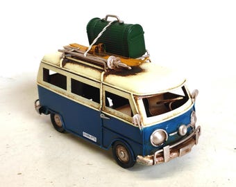 May include: A vintage-style blue and white van model with a luggage rack, skis, and a green suitcase on top. The van has a weathered appearance, suggesting a retro aesthetic. The model has a license plate with the number 22100-3772.