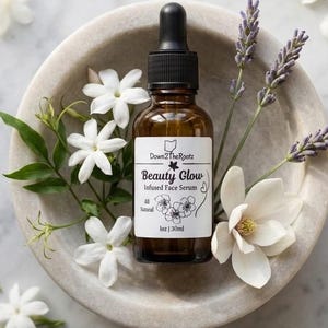 May include: A brown glass bottle of "Beauty Glow" infused face serum with a black dropper, surrounded by white flowers, green leaves, and lavender sprigs. The bottle is in a marble bowl. The label reads "All Natural" and "1 oz | 30ml".