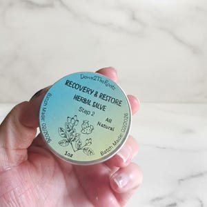 K&ouml;nnte beinhalten: Eine runde, silberfarbene Dose mit Kr&auml;utersalbe wird in einer Hand gehalten. Das Etikett lautet "RECOVERY & RESTORE HERBAL SALVE" mit dem Text "Step 2" und "All Natural". Die Dose enth&auml;lt 28 Gramm und hat ein Herstellungsdatum von 02/2026.