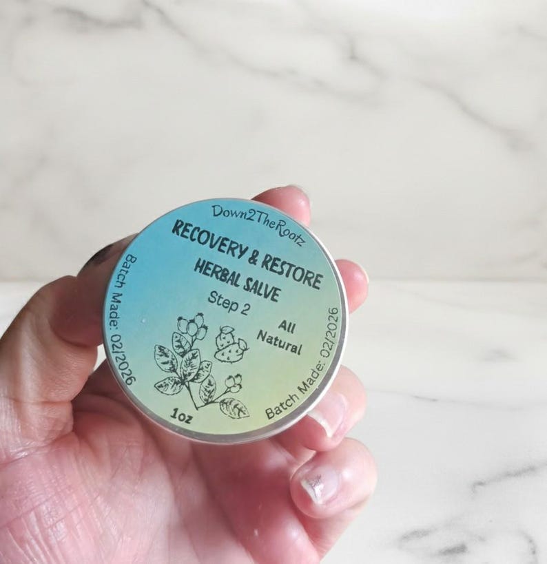 K&ouml;nnte beinhalten: Eine runde, silberfarbene Dose mit Kr&auml;utersalbe wird in einer Hand gehalten. Das Etikett lautet "RECOVERY & RESTORE HERBAL SALVE" mit dem Text "Step 2" und "All Natural". Die Dose enth&auml;lt 28 Gramm und hat ein Herstellungsdatum von 02/2026.