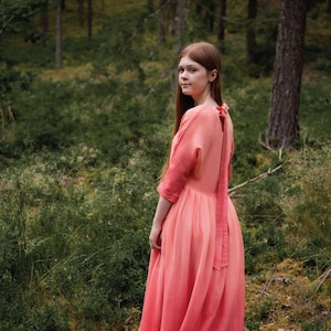Puede incluir: Vestido color coral con silueta fluida y detalle de lazo en la espalda. El vestido tiene mangas tres cuartos y una falda larga y con capas. La imagen está ambientada en un bosque con exuberante follaje verde y árboles altos.