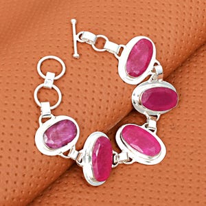 Könnte beinhalten: Ein silbernes Armband mit ovalen, tiefrosa Edelsteinen. Die Edelsteine sind in einzelnen Silberfassungen gefasst und durch Silberglieder verbunden. Das Armband hat einen Knebelverschluss. Die Edelsteine haben eine lebendige, gesättigte Farbe. Das Armband ist ca. 19 cm lang.