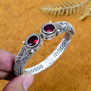 May include: A silver cuff bracelet featuring two oval-shaped, deep red garnet gemstones set in ornate, oxidised metalwork. The band displays intricate scroll patterns and a textured finish, with a hinged opening mechanism and a perforated inner design.