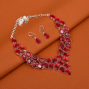 Può includere: Un set di collana e orecchini in argento. La collana e gli orecchini presentano più pietre preziose rosse a forma di goccia. I gioielli sono esposti su una superficie marrone.