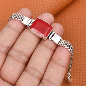 May include: A silver bracelet featuring a rectangular, faceted red stone set in a polished metal frame. The band is constructed from multiple thin, interconnected silver chains that attach to the central stone setting on both sides.