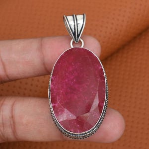 Puede incluir: Colgante ovalado con una gran piedra preciosa de rubí facetada. El rubí es de color rojo intenso y está engastado en un marco de metal plateado. El colgante tiene una argolla decorativa para sujetar una cadena.