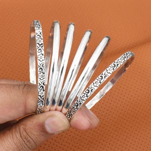 May include: A collection of silver bangles. Some bangles have a floral pattern, while others are plain. The bangles are displayed against a warm, orange-brown background. The jewelry is held in a hand.