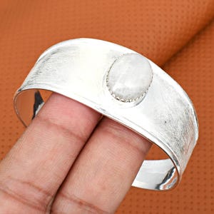 May include: A wide, silver cuff bracelet with a textured finish. The bracelet features an oval, off-white gemstone set in the centre. The bracelet is held by a hand against a brown background.