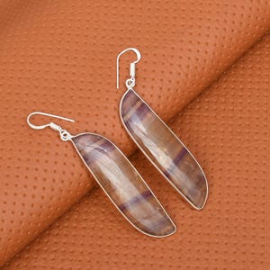 May include: A pair of silver-toned earrings with elongated, curved, rectangular stones. The stones have a translucent, striped appearance with shades of brown and purple. The earrings are displayed on a textured, brown surface.