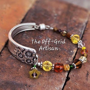 May include: A handcrafted bracelet featuring a repurposed silver spoon as a focal point. The spoon's handle is adorned with floral details. The bracelet incorporates colorful beads in shades of yellow, orange, and brown, with a star charm. The text "The Off-Grid Artisan" is visible.