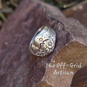May include: A silver ring with floral and dot details. The ring has a spoon-like shape with a rounded top and a detailed floral design. The text "The Off-Grid Artisan" is visible in the image.