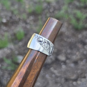 May include: A silver-toned ring featuring an embossed floral pattern with a central rose bud and surrounding leaves. The band has a wide, flat profile and is displayed on a tapered, polished brown wooden mandrel, showcasing the intricate metalwork.