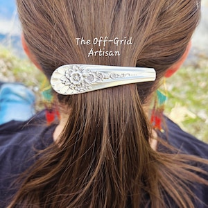 May include: A silver-toned metal hair clip crafted from a repurposed spoon handle, featuring an embossed floral pattern. The text The Off-Grid Artisan is visible above the clip. The accessory is secured in brown hair, holding a portion of the hair back.
